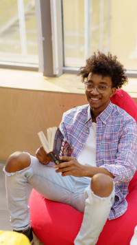 A high school student sits on a bean bag chair, holding a book and smiling.
