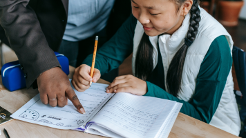 A student looks at a worksheet. A teacher points to the worksheet. The student smiles, focused.