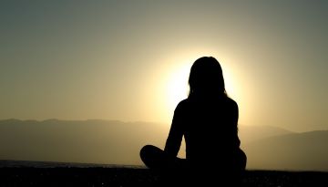 A person meditates at dusk. The photo captures their silhouette with the sun setting in the background.