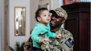 A person in military camouflage holds a toddler. They are both smiling.