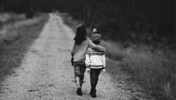 A black and white photo of two siblings walking along a dirt road. The taller sibling has their arm around the shorter sibling's shoulders.