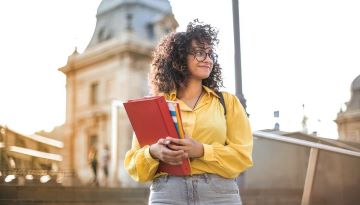 [ID: A young adult holds binders and notebooks as they stand outside at what appears to be a college campus. It is a bright sunny day. The person looks in the distance and smiles.]